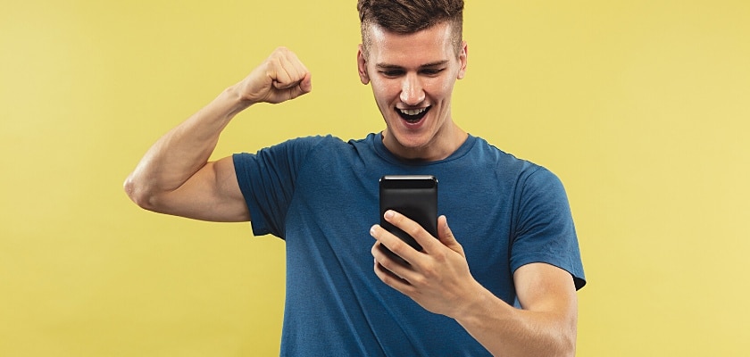 Primo piano frontale di un ragazzo che fa il gesto della vittoria con il braccio destro mentre guarda il touchscreen del suo iPhone – Matched Betting