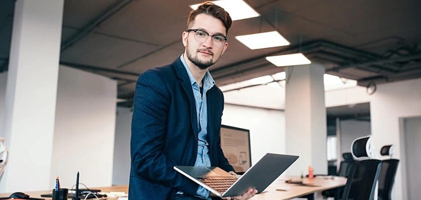 Uomo d’affari con gli occhiali seduto vicino al posto di lavoro in ufficio che tiene un laptop e guarda la fotocamera