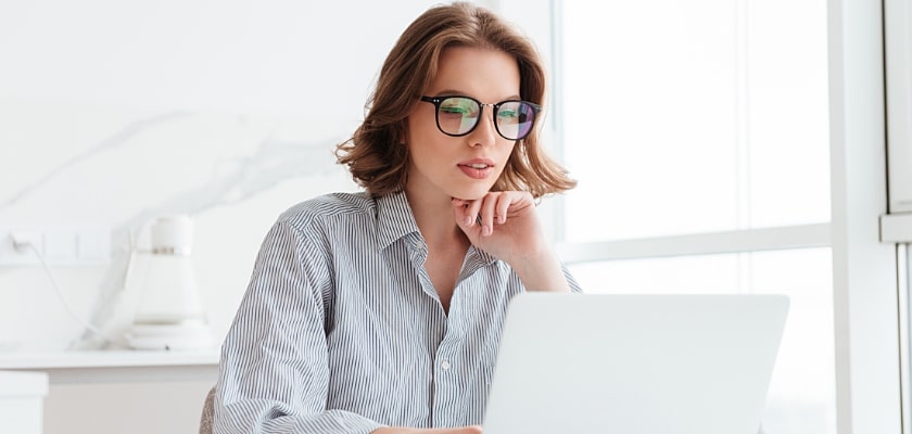 Affascinante donna d’affari con gli occhiali con la camicia a righe che lavora con il computer portatile mentre si trova a casa